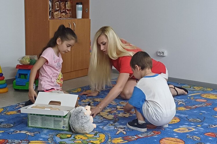 Trained staff at Kherson’s Resilience Center kneel beside children on an illustrated space-themed rug. Intentional care includes non-threatening, engaging toys and adults close enough to offer safety, but never crowding.