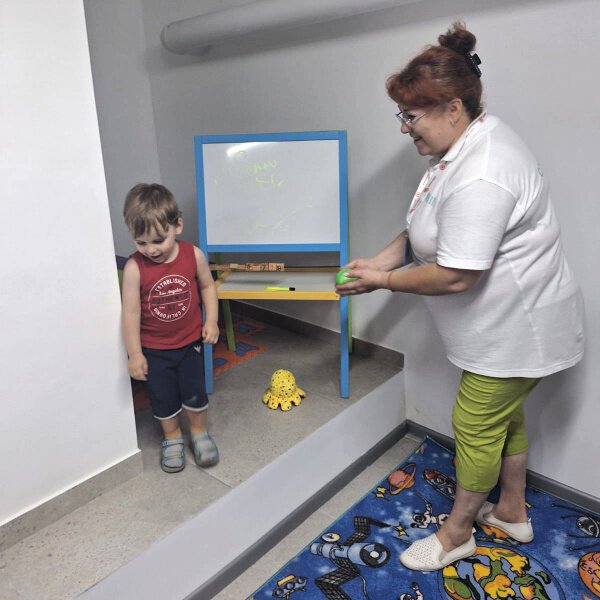 Ms. Anna Samchuk gently engages a small boy in play. The Center is clean, contained, safe, and designed for children navigating stress responses and interrupted development. She is not simply supervising; she is co-regulating.
